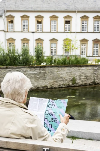 Eine ältere Person blickt in ein Programmheft zur Ausstellung, im Hintergrund die Pader und die Stadtbibliothek.