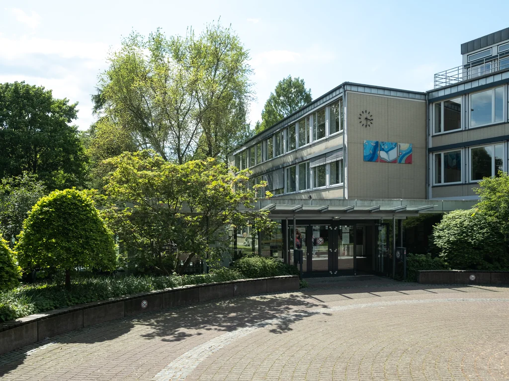 The image depicts the entrance of a modern building with large windows and a clock on the facade, surrounded by lush green trees and bushes. A paved path leads to the main entrance, bordered by well-maintained plants. The building  is the Westfalenkolleg in Paderborn.
