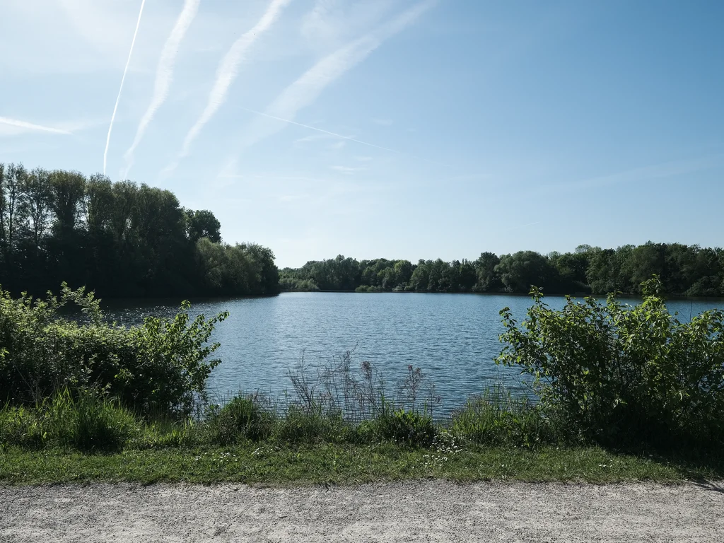 The view shows the calm, blue shimmering Pader lake, framed by dense greenery along the shore. The sky is clear with white contrails stretching across it.