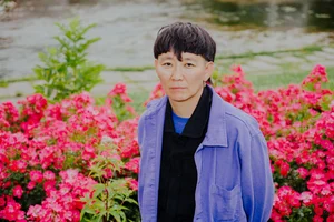 English image description:
A person with short dark hair stands calmly in front of a bed of bright pink flowers, with flowing water in the background. They wear a blue jacket over dark clothing and look directly at the camera.