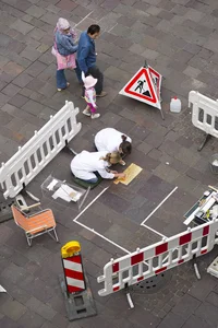 The two artists kneel on the ground and install golden plates on the path. A family watches them go by.