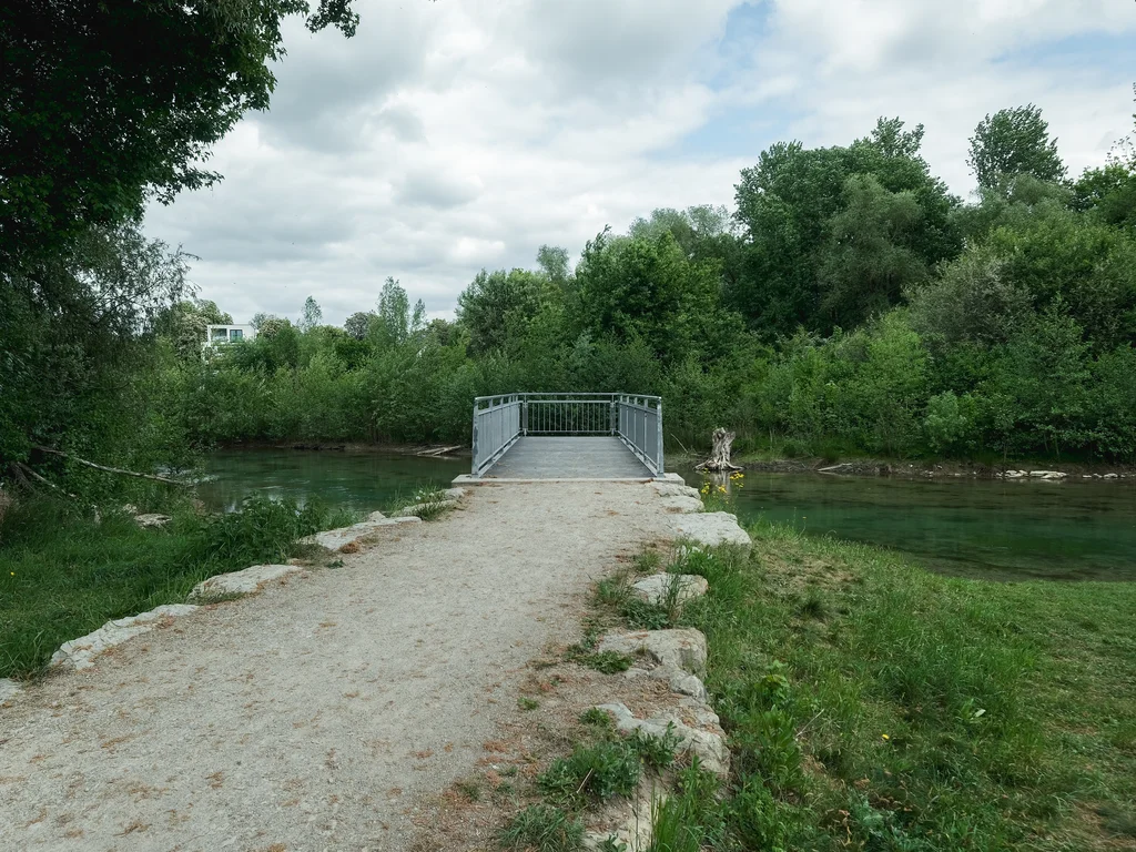 The picture shows a narrow metal viewing platform on the river Pader, surrounded by lush green vegetation and trees. A sandy path leads to the platform, which is lined with rocks and green plants.