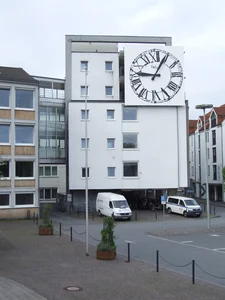 The picture shows a modern white building with a large round clock face on the façade. In front of the building are two white vans and some bicycles, and the area is landscaped with plants and bollards.