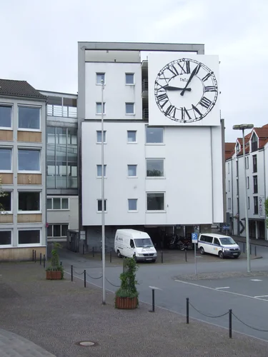 The picture shows a modern white building with a large round clock face on the façade. In front of the building are two white vans and some bicycles, and the area is landscaped with plants and bollards.
