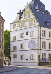 The picture shows a historic building with a white façade and golden accents, which stands out due to its elaborate decorations and a large, artistic mural with a peacock motif.