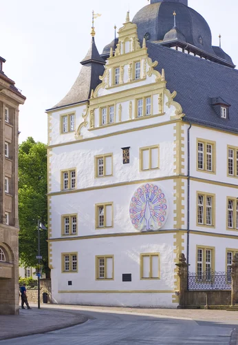 The picture shows a historic building with a white façade and golden accents, which stands out due to its elaborate decorations and a large, artistic mural with a peacock motif.