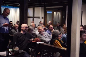 Das Bild zeigt eine Gruppe von Menschen, die in einem Raum mit großen Fenstern und Vorhängen an einem runden Tisch sitzen. Die Personen sind in Gespräche vertieft, und auf dem Tisch stehen Getränke und Snacks.