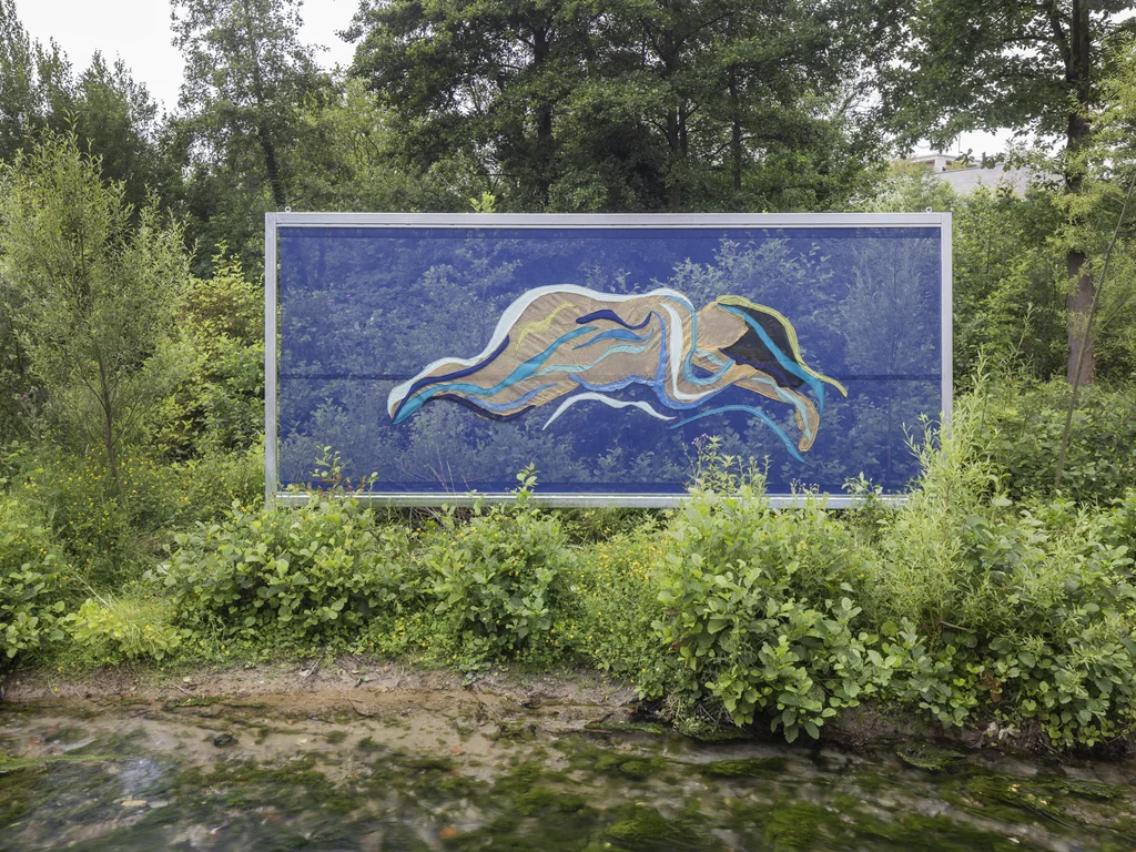 A large-format poster displays an abstract, wave-like figure in shades of blue, beige, and green, set amidst dense greenery by the water. The figure appears to float over a vivid blue background that reflects the sky and surrounding trees.