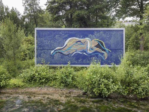A large-format poster displays an abstract, wave-like figure in shades of blue, beige, and green, set amidst dense greenery by the water. The figure appears to float over a vivid blue background that reflects the sky and surrounding trees.