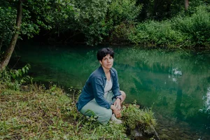 Eine Person hockt am Ufer eines ruhigen, grün schimmernden Gewässers, umgeben von dichtem Wald und üppiger Vegetation. Die Szene wirkt still und naturverbunden.