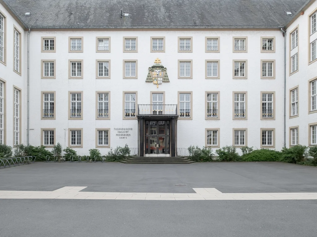 The image depicts the front of a historic building with symmetrically arranged windows and a central entrance. Above the entrance, there is a coat of arms and the inscription "Theologische Fakultät Paderborn."