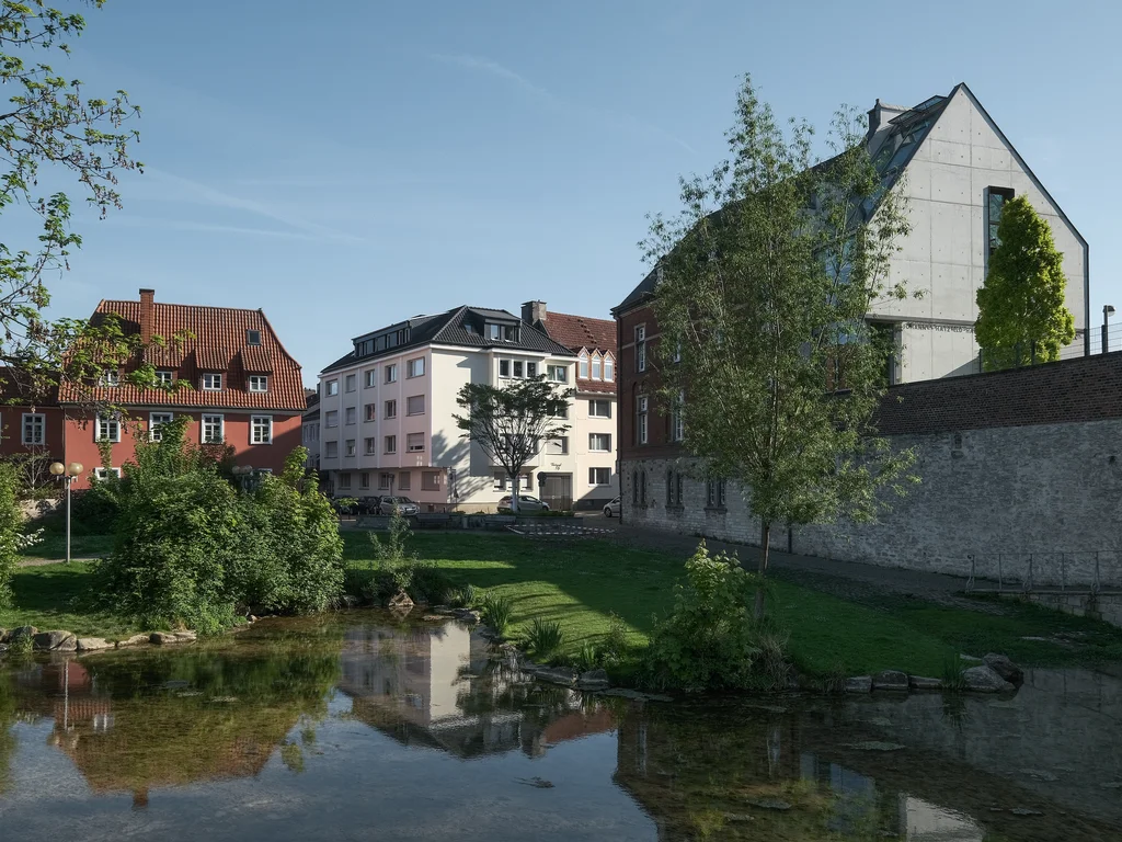 The image depicts a tranquil scene with a small river, the Dielenpader, flowing through a green landscape, surrounded by trees and bushes. In the background, there are various buildings, including a modern house with a glass facade and traditional houses with red roofs.
