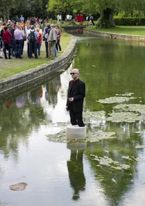 A man in a black suit with a red bow tie and sunglasses stands on a small round platform in the middle of a shallow body of water. On the shore, a large group of people are watching the scene.
