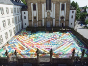 The picture shows a large, colorful square with a geometric pattern of colorful tiles surrounded by a wrought iron railing. In the background is a historic building with high windows and a classical façade, Paderborn Cathedral.