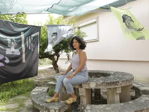 Eine Frau mit schulterlangen dunklen Haaren sitzt auf einer runden Steintisch-Bank-Kombination im Freien. Um sie herum hängen kunstvoll gestaltete Stoffbahnen mit applizierten Figuren unter einem grünen Sonnenschutz. Bei der Frau handelt es sich um die Textilkünstlerin Sophie Utikal.