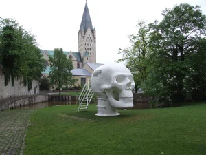 The picture shows a large white sculpture of a skull with a ladder leading into the skull's mouth. In the background is a church with a high tower, Paderborn Cathedral, surrounded by trees and a small river, the Pader.