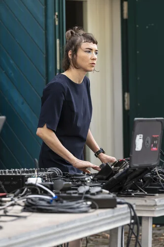 Eine Person steht konzentriert hinter einem Tisch voller Audiotechnik und Laptop und mischt Musik; sie trägt ein dunkelblaues Kleid und hat markantes Augen-Make-up. Es ist die Künsterin Jana Kerima. Der Hintergrund zeigt eine geöffnete Tür und eine dunkelgrüne Holzwand.