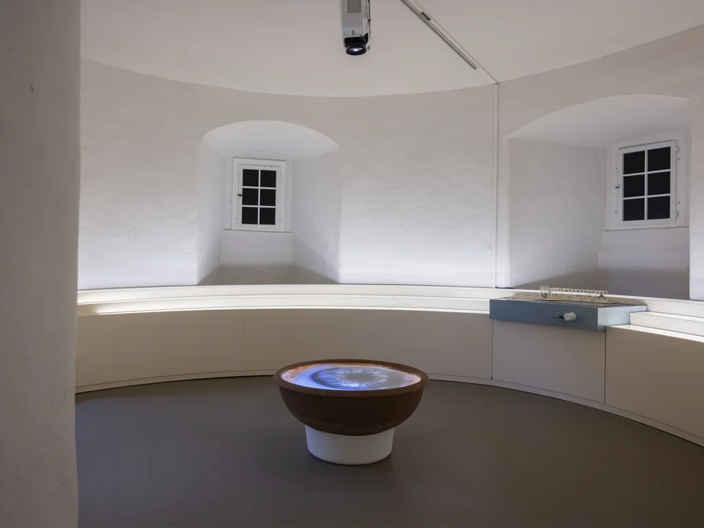 A bright, circular room with white plastered walls features a projection of an eyeball in a bowl-shaped installation at its center. The space is surrounded by a white bench and a small architectural model displayed on a pedestal.