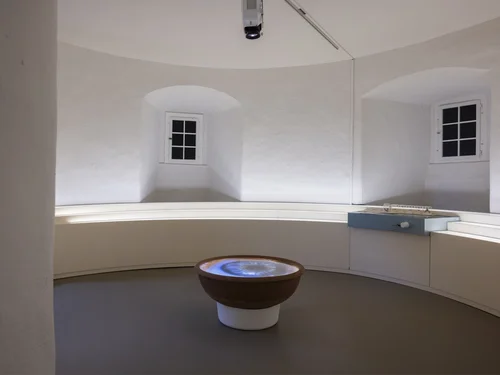 Ein heller, runder Raum mit weiß verputzten Wänden zeigt in der Mitte eine Projektion eines Augapfels in einer schalenartigen Installation. Umgeben ist der Raum von einer weißen Sitzbank und einem kleinen Architekturmodell auf einem Podest.