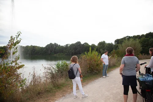 Eine Gruppe schaut einem Mann zu, der ein Gedicht vorliest. Er steht vor der Wasserfontäne, die Teil ist von Jeppe Heins Installation am Padersee.