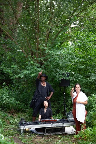 Das Bild zeigt drei Personen in einem grünen Wald: Eine Person sitzt an einem Tisch mit elektronischem Musik-Equipment, eine weitere steht daneben und deutet nach oben, während die dritte Person mit ausgestrecktem Arm in die Kamera zeigt.