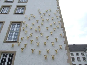 The picture shows the façade of a building decorated with numerous golden angel figures arranged in a regular pattern. The angels are attached to the wall and have wings, with some of them holding small objects or instruments.