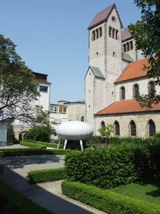 The picture shows a historic church with a high tower and red roof tiles, surrounded by a well-tended garden with clipped hedges. In the foreground is a modern, white sculpture that looks like a futuristic object on legs, creating a contrast to the traditional architecture.