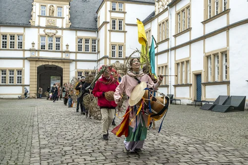 Eine bunt gekleidete Gruppe zieht trommelnd und mit großen, geflochtenen Weidenobjekten durch einen historischen Innenhof. Angeführt wird der Umzug von einer Performerin mit Trommel und wehenden Stoffbändern.