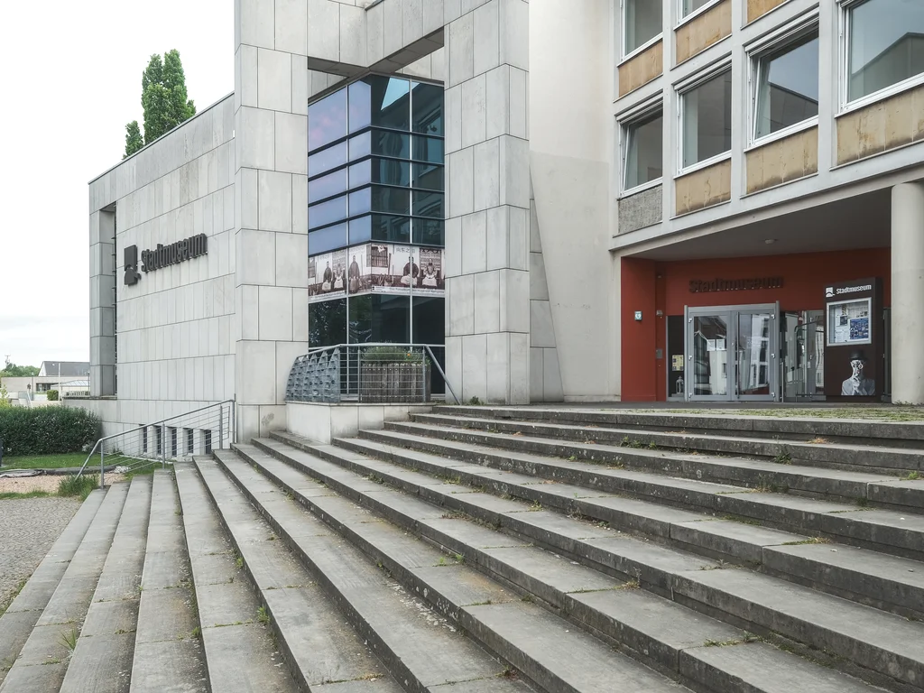 The image depicts the entrance of a modern museum building with a wide staircase leading to the main entrance. The building has a smooth, gray facade with large glass windows and the inscription "Stadtmuseum."