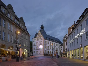 The picture shows an evening cityscape with an illuminated street surrounded by historic and modern buildings. In the center is a striking white building with an ornate façade and a dark roof with turrets. A colorful, round light pattern shines on its wall, reminiscent of a church window or a modern light installation. The luminous pattern on the wall of the white building depicts a peacock with an open wheel. It is a work of art by Silke Wagner entitled “Sometimes” for the exhibition Tatort Paderborn 2014 and consists of colorful neon tubes that trace the shape of the majestic bird. The peacock's body is placed in the center, while the artfully curved tail feathers radiate behind it in a large, fan-like pattern. It is the artwork by Silke Wagner entitled “Sometimes” for the exhibition Tatort Paderborn 2014.