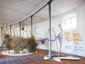 Das Bild zeigt einen Ausstellungsraum mit großen, kunsthandwerklichen Neststrukturen aus Weiden-Zweigen, sowie mit handschriftlichen Texten an der Wand auf Deutsch und Englisch.