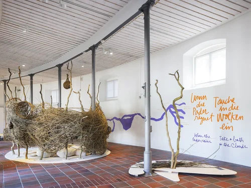 The image depicts an exhibition space with large, handcrafted nest structures made of branches, along with handwritten texts on the wall in German and English.