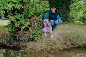 Zwei Personen hocken nebeneinander am Ufer der Pader, neben einem großen, verwurzelten Baum. Umgeben von dichtem Grün blicken sie gedankenverloren in verschiedene Richtungen. Es sind der Künstler Lex Rütten und die Künstlerin Jana Kerima Stolzer.