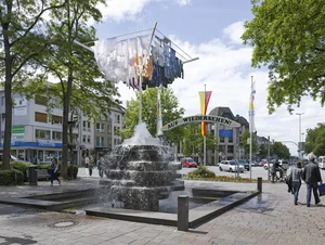 The picture shows a lively square with a central fountain, above which hovers an artistic installation consisting of a large clothesline with clothes hanging in it.