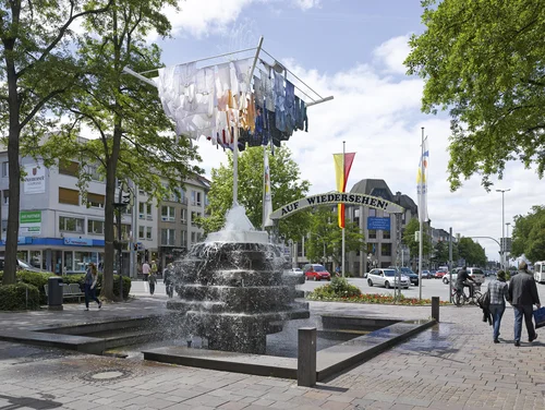 The picture shows a lively square with a central fountain, above which hovers an artistic installation consisting of a large clothesline with clothes hanging in it.