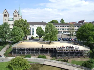 The picture shows a large, open area with a group of people gathered on a raised platform, surrounded by a metal railing. A church with a green tower, modern buildings and trees belonging to the city of Paderborn can be seen in the background.