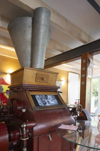 Eine große, alte rote Industriemaschine steht in einem warm beleuchteten Innenraum; in ihrem mittleren Bereich ist ein Bildschirm eingelassen, der eine schwarz-weiße Flusslandschaft zeigt. Oben ragen zwei große trichterförmige Metallaufsätze empor, und davor steht ein runder Glastisch mit Papieren.