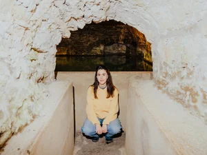 Das Bild zeigt eine junge, hockende Frau mit langem, dunkelbraunem Haar, die einen hellgelben, gemusterten Pullover trägt. Sie scheint sich in einem Keller zu befinden und hat einen neutralen Gesichtsausdruck.