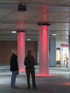 The picture shows two people standing under a covered area with illuminated red pillars. A store in paderborn city center can be seen in the background, and the scene has a modern and urban feel.