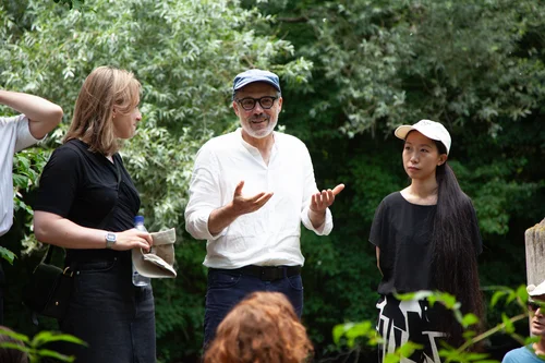 Das Bild zeigt drei Personen im Freien vor grünem Blätterhintergrund: Eine Person in Weiß mit Basecap und Brille spricht gestikulierend, während zwei weitere Personen aufmerksam zuhören.