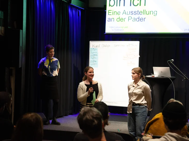 Zwei Referentinnen stehen vor der Bühne vor einem Whiteboard mit den Wörtern "Kunst", "Ökologie", "Geschichte", und "Städtische Entwicklung".