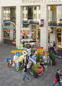 The picture shows a lively street scene in front of a store with a variety of traffic and advertising signs, including an eye-catching stop sign, place name signs and an artistically designed horse. People walk by, while the stores of Paderborn city center can be seen in the background.