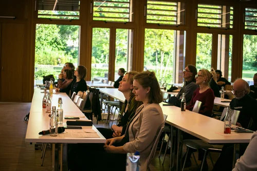 Several people are sitting at tables arranged in rows. They are all facing forward and appear to be listening attentively.