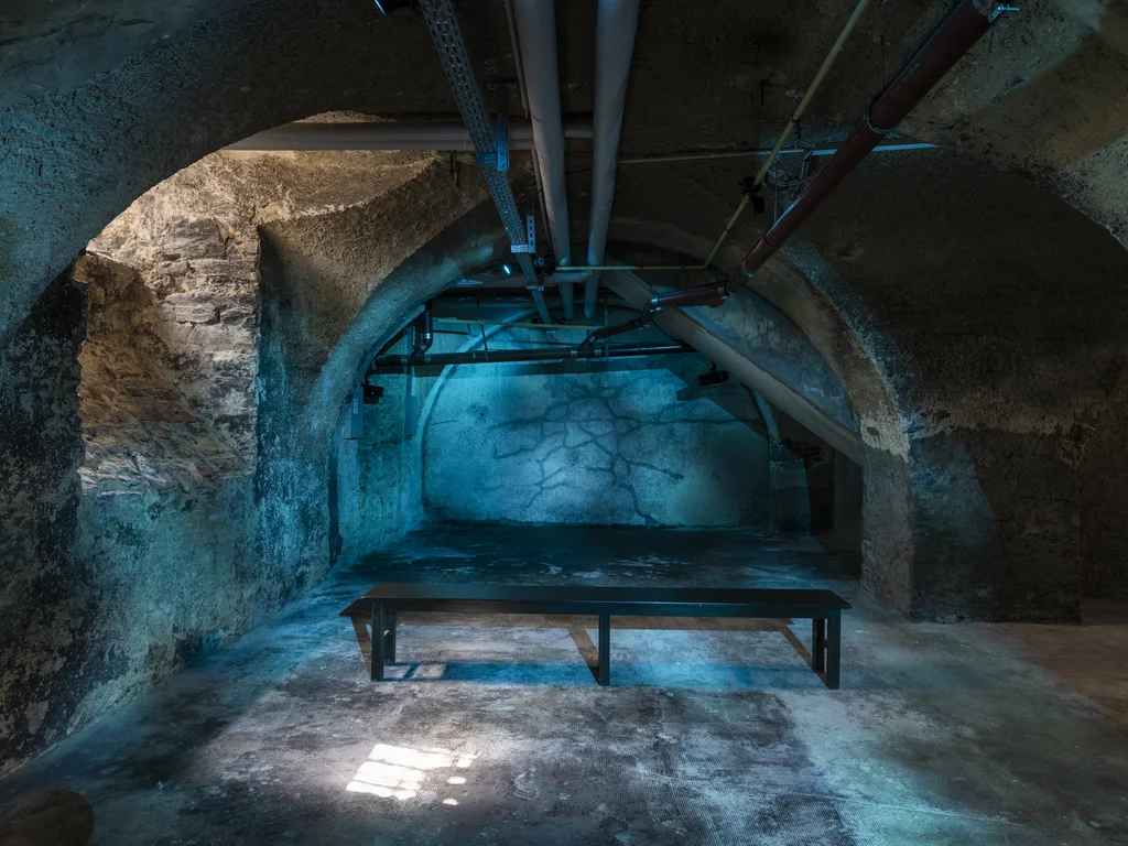 Ein alter, gewölbter Kellerraum aus Stein ist in kühles blau-türkises Licht getaucht; in der Mitte steht eine einfache schwarze Bank vor einer Wand mit rissigen Strukturen. Über Kopf verlaufen sichtbare Rohre.
