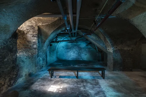 An old vaulted stone cellar is bathed in cool blue-turquoise light; in the center stands a simple black bench facing a wall marked by branching cracks. Overhead you see exposed pipes.