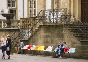 A row of chairs in different colors stand in front of an old building. People are sitting on some of the chairs, others are walking past.