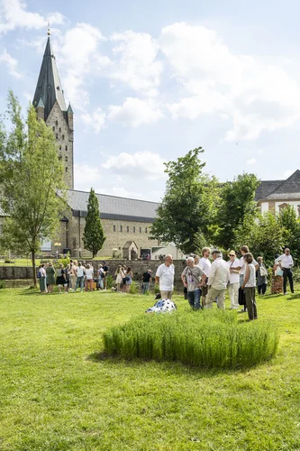 Mehrere Gruppen von Menschen stehen und unterhalten sich auf einer großen Wiese, im Hintergrund ragt der Turm des Paderborner Doms auf. In der Mitte ist ein rundes Stück höher wachsendes Flachs zu sehen, das wie eine kleine Installation wirkt.