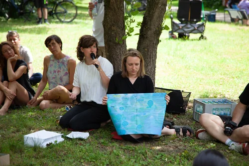 Das Bild zeigt eine Gruppe von Menschen, die im Grünen auf dem Rasen sitzen; eine Person hält ein Mikrofon, während eine andere ein großes, blaues Plakat präsentiert.