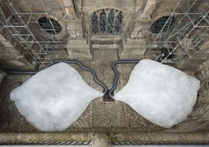 The picture shows a modern art installation on a historic building, consisting of two large, white, organically shaped structures connected by tubes. The installation incorporates the scaffolding surrounding the building.