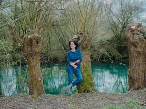 Das Bild zeigt eine Frau mit dunkelbraunem Haar, die ein dunkelblaues Oberteil trägt und vor drei Weiden am Fluss, der Pader, steht. Sie trägt große, grüne Ohrringe und lächelt sanft. Bei der Frau handelt es sich um die Künstlerin Daniela Brasil.